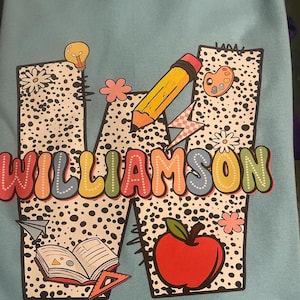 May include: A blue shirt with a large white letter W with black polka dots. The letter W is decorated with a lightbulb, a pencil, a paint palette, a book, a ruler, an apple, and the word "WILLIAMSON" in colorful letters.