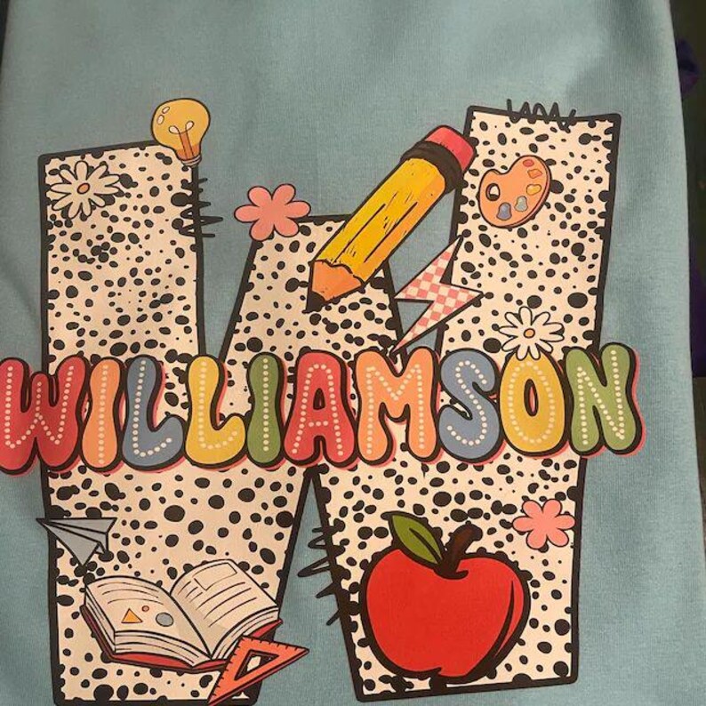 May include: A blue shirt with a large white letter W with black polka dots. The letter W is decorated with a lightbulb, a pencil, a paint palette, a book, a ruler, an apple, and the word "WILLIAMSON" in colorful letters.