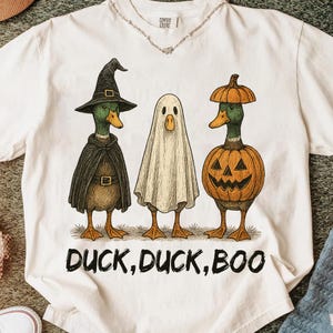 May include: White t-shirt featuring three ducks dressed in Halloween costumes. One duck is in a witch costume, another is a ghost, and the third is in a pumpkin costume. The text "DUCK, DUCK, BOO" is printed below the ducks.