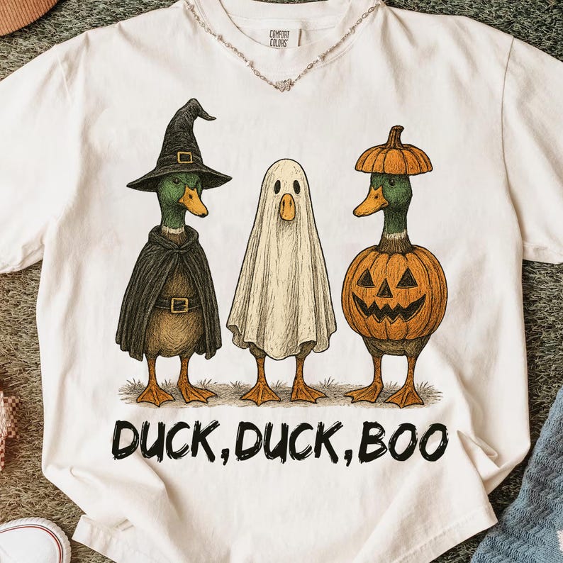 May include: White t-shirt featuring three ducks dressed in Halloween costumes. One duck is in a witch costume, another is a ghost, and the third is in a pumpkin costume. The text "DUCK, DUCK, BOO" is printed below the ducks.