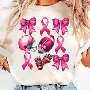 May include: Cream-colored t-shirt featuring a collection of pink football-themed graphics. The design includes pink ribbons, a football, a helmet, and gloves. The graphics are arranged in a grid pattern.