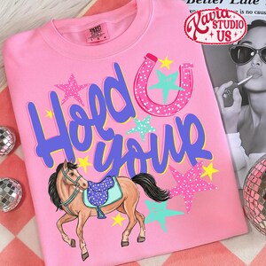 May include: Pink t-shirt with the phrase "Hold Your Horses" in blue and a horseshoe and stars design. A cartoon horse with a saddle is featured below the text. The shirt has a crew neck and a tag on the upper left.