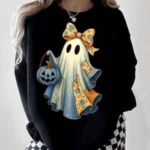 Op de afbeelding: Zwart sweatshirt met een grillige spookillustratie. Het spook is versierd met een bloemenstrik en patchworkdetails, met een blauwe pompoenvormige snoepzak. Een charmant Halloween-ontwerp.