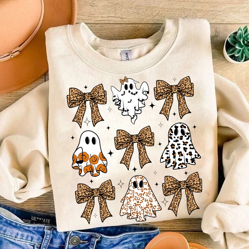 May include: Cream-colored sweatshirt featuring a Halloween-themed design. The design includes several cartoon ghosts with bows and star accents. The bows have a leopard print pattern. The ghosts have different patterns and expressions.