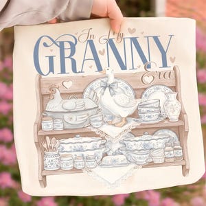 May include: A cream-colored decorative pillow featuring a watercolor illustration of a wooden shelf filled with vintage-style blue and white china, a goose, and a chicken. The text "In My Granny Era" is written across the top in blue script.
