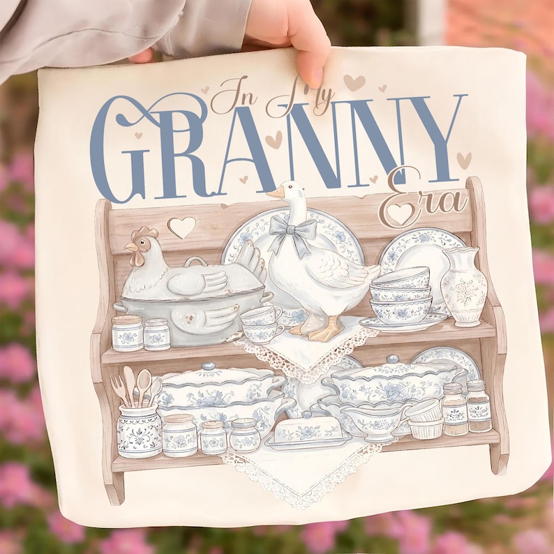 May include: A cream-colored decorative pillow featuring a watercolor illustration of a wooden shelf filled with vintage-style blue and white china, a goose, and a chicken. The text "In My Granny Era" is written across the top in blue script.