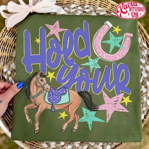 May include: Olive green t-shirt with the words "Hold Your Horses" in purple and yellow. A cartoon horse with a saddle is featured below the text, along with stars and a horseshoe design.