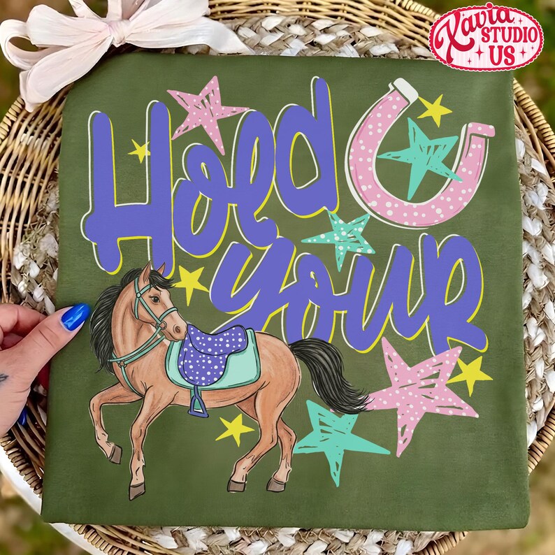 May include: Olive green t-shirt with the words "Hold Your Horses" in purple and yellow. A cartoon horse with a saddle is featured below the text, along with stars and a horseshoe design.