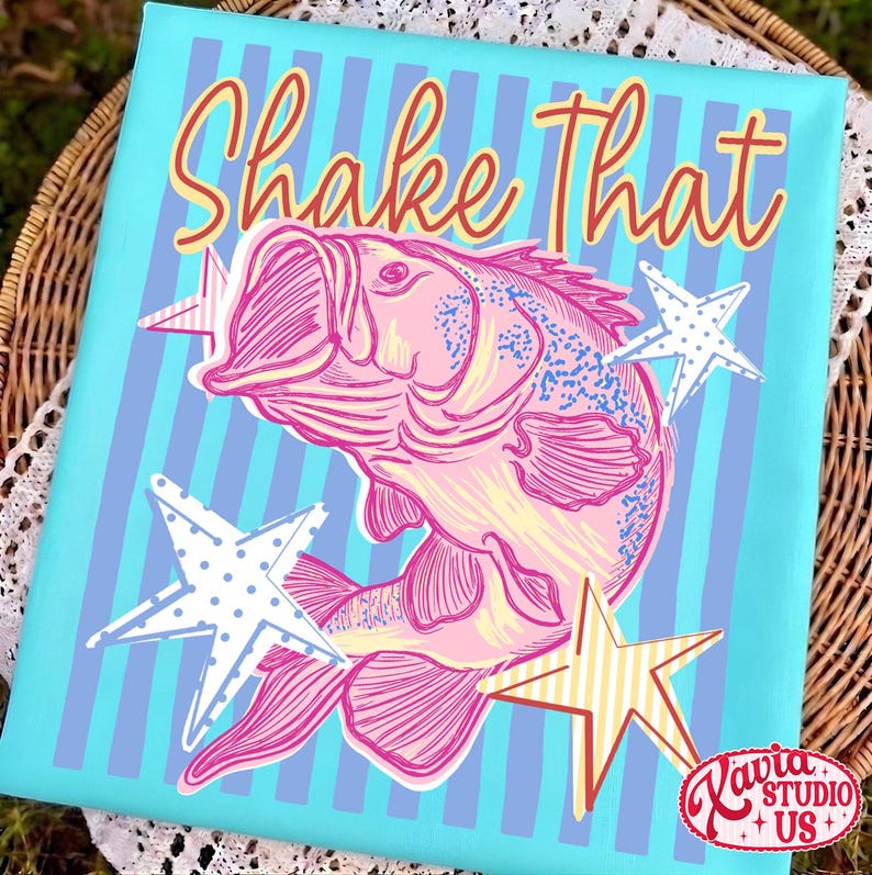 May include: A canvas art print featuring a pink fish illustration with the text "Shake That" in a playful font. The background is light blue with vertical stripes, and the design includes star accents. The artwork is signed "Ravia Studio US".