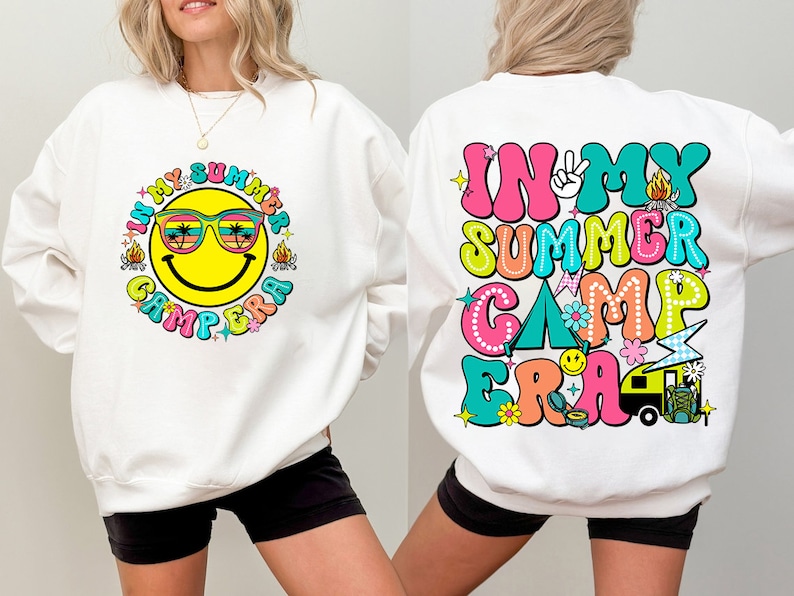 May include: White sweatshirt with a colorful graphic design. The front features a smiling face wearing sunglasses with the text "IN MY SUMMER CAMP ERA". The back of the sweatshirt has the same text in a retro font with camping-themed illustrations.