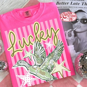 May include: A hot pink t-shirt with the word "Lucky" in a light green script. The shirt features a graphic of a duck with outstretched wings, surrounded by stars. The shirt is on a pink and white checkered surface.
