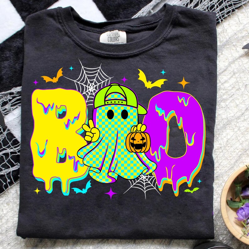 Puede incluir: Camiseta gris oscuro con un dise&ntilde;o colorido de Halloween. La palabra "BOO" est&aacute; escrita en letras goteantes amarillas y moradas. Un fantasma de dibujos animados con una gorra verde y una calabaza est&aacute; en el centro, con murci&eacute;lagos, estrellas y telara&ntilde;as.