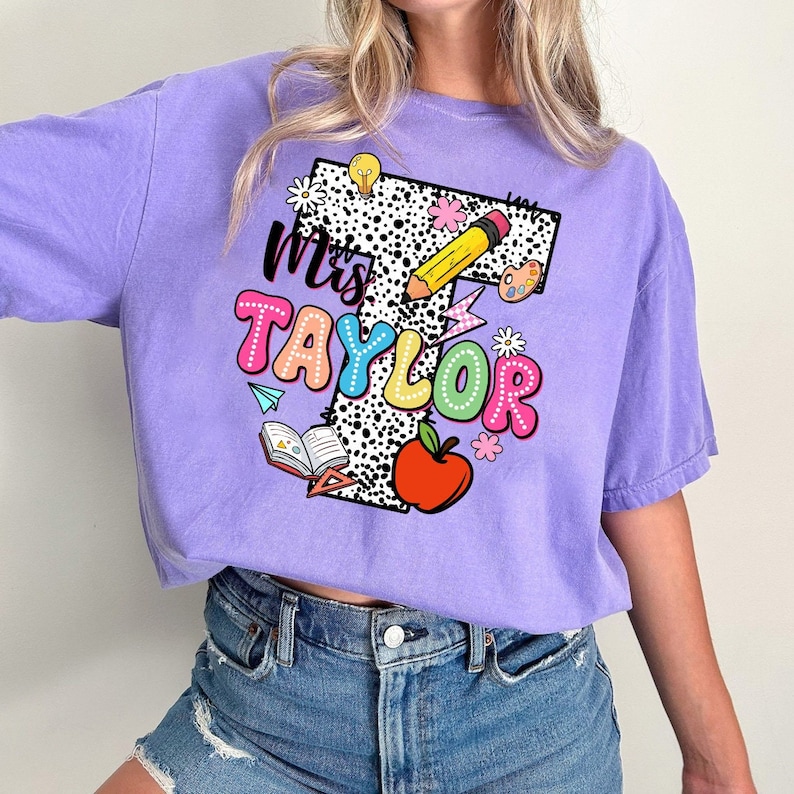 May include: Purple t-shirt with a white and black polka dot letter T. The letter T is decorated with colorful school supplies, including a pencil, an apple, a book, and a lightbulb. The text "Mrs. Taylor" is written in rainbow letters.