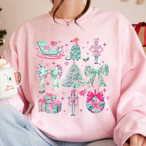 May include: A light pink sweatshirt featuring a festive Christmas design. The design includes a sleigh, a rat in a coat, a nutcracker, bows, a Christmas tree, wrapped gifts, and a Christmas ornament. The illustrations are in shades of pink, green, and white.