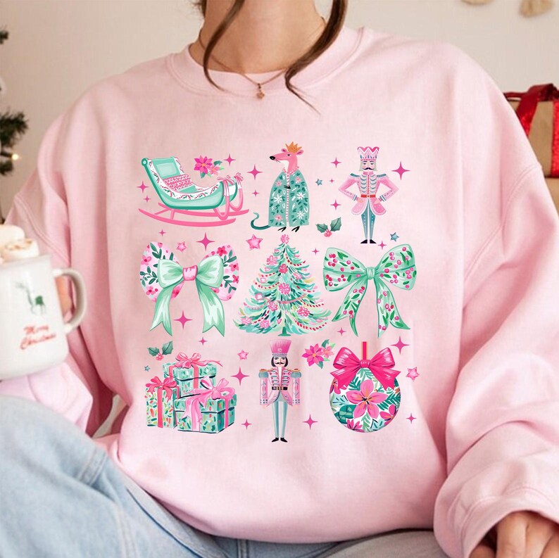 May include: A light pink sweatshirt featuring a festive Christmas design. The design includes a sleigh, a rat in a coat, a nutcracker, bows, a Christmas tree, wrapped gifts, and a Christmas ornament. The illustrations are in shades of pink, green, and white.