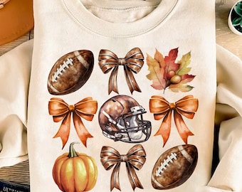 May include: Cream-coloured sweatshirt with a watercolour design of a football, football helmet, pumpkin, bows, and autumn leaves. The design is in shades of brown, orange, and yellow. A perfect autumn or football season themed top.