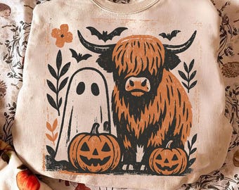 May include: Beige sweatshirt featuring a Halloween-themed graphic. The design includes a Highland cow, a ghost, two jack-o'-lanterns, bats, and floral elements in black and orange. The sweatshirt has a crew neck and long sleeves.