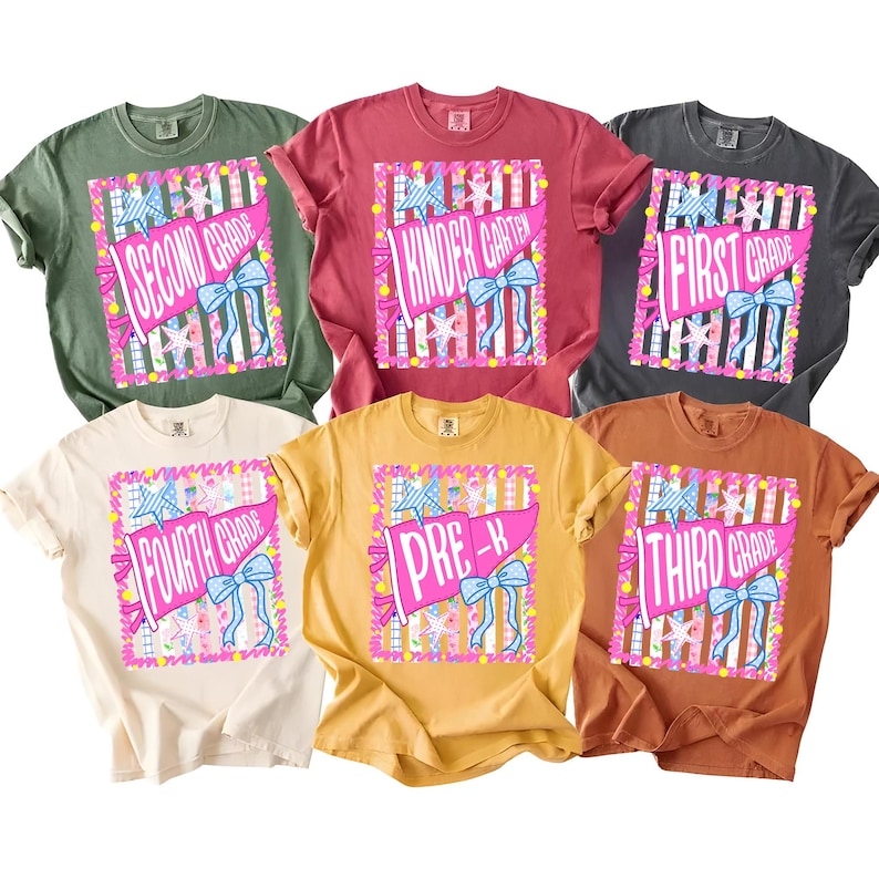 May include: Six short-sleeve crew neck t-shirts in olive green, red, charcoal gray, cream, mustard yellow, and terracotta. Each shirt features a graphic print of a pink pennant flag with a blue bow, stars, and text: Second Grade, Kindergarten, First Grade, Fourth Grade, Pre-K, and Third Grade.