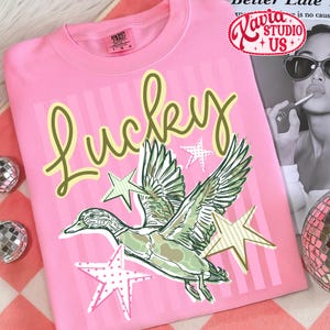 May include: Pink t-shirt with the word "Lucky" in a script font, a graphic of a duck with outstretched wings, and star accents. The shirt has a crew neck and short sleeves.
