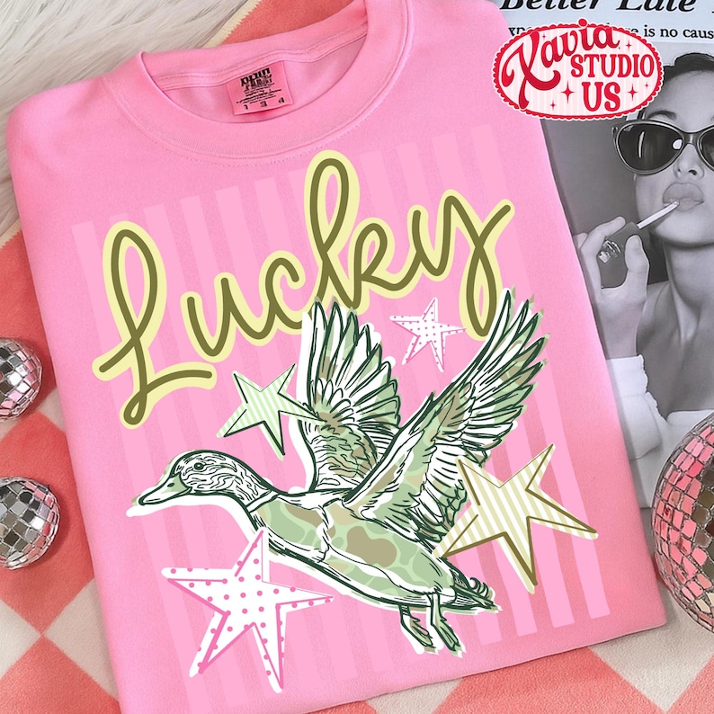 May include: Pink t-shirt with the word "Lucky" in a script font, a graphic of a duck with outstretched wings, and star accents. The shirt has a crew neck and short sleeves.