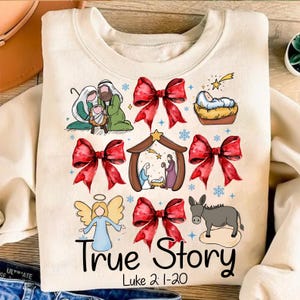 May include: Cream-colored sweatshirt with a colorful Christmas-themed graphic. The design includes illustrations of the Nativity scene, red bows, an angel, and a donkey. The text "True Story" and "Luke 2:1-20" are printed below the images.