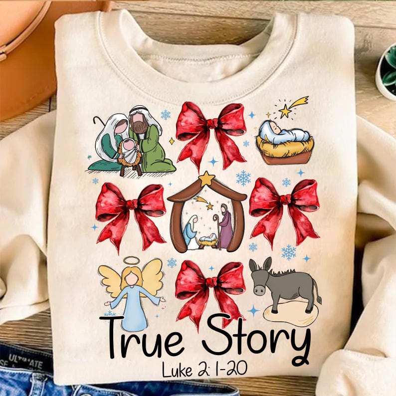 May include: Cream-colored sweatshirt with a colorful Christmas-themed graphic. The design includes illustrations of the Nativity scene, red bows, an angel, and a donkey. The text "True Story" and "Luke 2:1-20" are printed below the images.