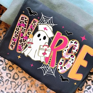 May include: Dark gray sweatshirt with the word "NURSE" in pink, leopard print, and gold lettering. A cartoon ghost in a nurse's hat and carrying a medical bag is in the center. Bats and spiderwebs decorate the design.