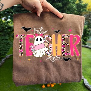 Op de afbeelding: Een bruin stoffen item met het woord "TEACHER" in roze en oranje, versierd met een spook dat een potlood vasthoudt, vleermuizen en spinnenwebben. De letter "T" heeft een luipaardprint.