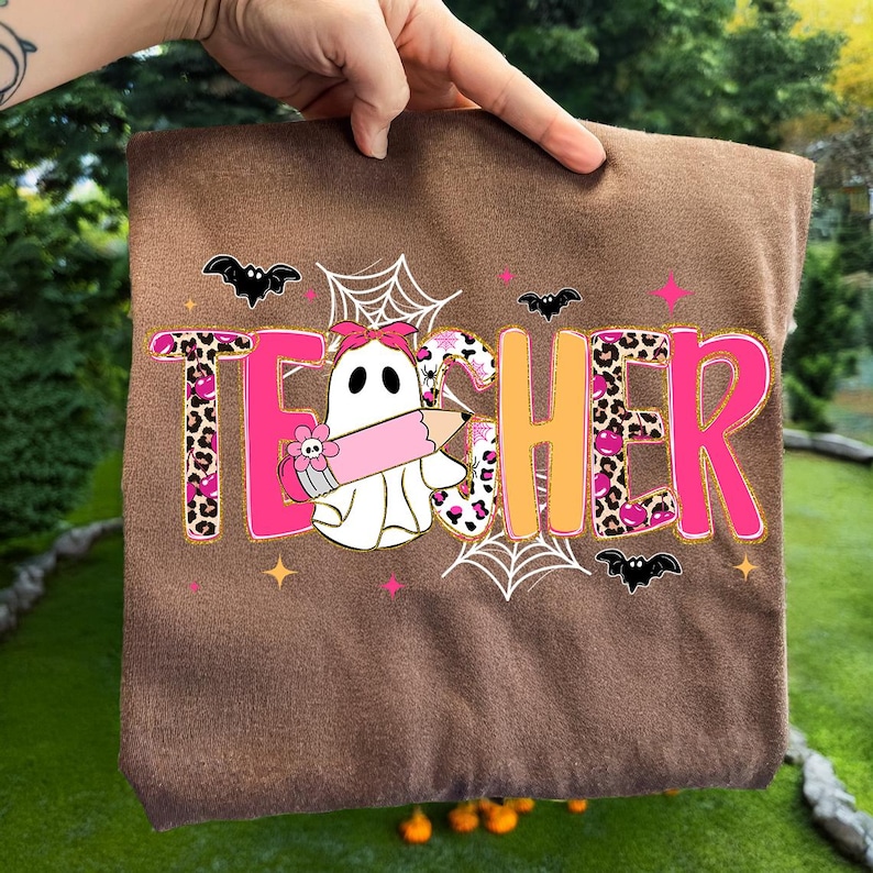 Op de afbeelding: Een bruin stoffen item met het woord "TEACHER" in roze en oranje, versierd met een spook dat een potlood vasthoudt, vleermuizen en spinnenwebben. De letter "T" heeft een luipaardprint.