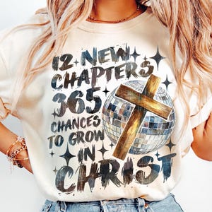 May include: Cream-colored sweatshirt with the text "12 New Chapters 365 Chances to Grow in Christ." The design includes a wooden cross over a disco ball and star accents. The shirt is a casual, faith-based apparel item.