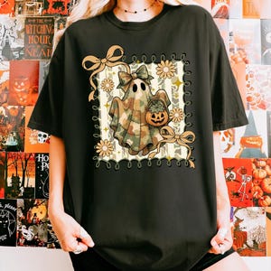 May include: Black t-shirt featuring a Halloween-themed graphic. The design includes a ghost wearing a camouflage sheet, holding a pumpkin basket, and adorned with bows and flowers. The graphic is framed with a decorative border.
