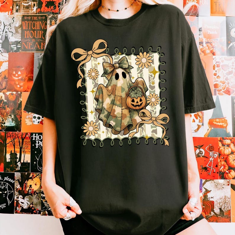 May include: Black t-shirt featuring a Halloween-themed graphic. The design includes a ghost wearing a camouflage sheet, holding a pumpkin basket, and adorned with bows and flowers. The graphic is framed with a decorative border.