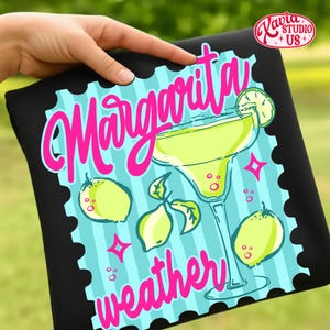 May include: Black t-shirt with a margarita design. The design features a margarita glass, lemons, and the words "Margarita weather" in pink script. The background is light blue with vertical stripes.