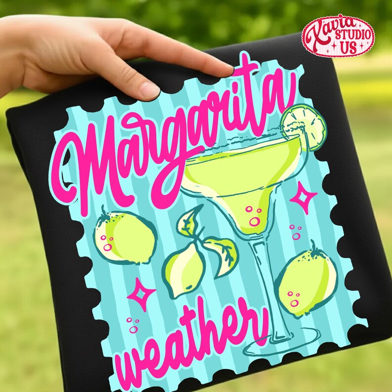 May include: Black t-shirt with a margarita design. The design features a margarita glass, lemons, and the words "Margarita weather" in pink script. The background is light blue with vertical stripes.