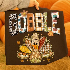 May include: A brown Thanksgiving-themed graphic featuring the word "GOBBLE" in a patchwork design with various patterns. Below, a cartoon turkey with patterned feathers and a polka-dot body is displayed. The design includes fall leaves and stars.