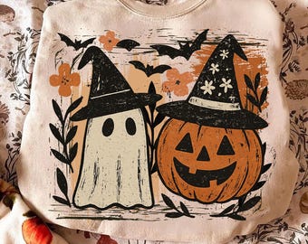 May include: Beige Halloween-themed sweatshirt featuring a ghost and a jack-o'-lantern, both wearing witch hats. The design includes bats, flowers, and foliage in black, orange, and beige. The sweatshirt is a crew neck style.