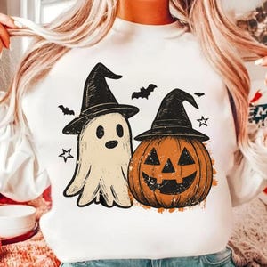 Op de afbeelding: Wit sweatshirt met een Halloween-ontwerp. Het ontwerp bevat een spook met een heksenhoed, een pompoen met een heksenhoed, vleermuizen en sterren. Het spook is gebroken wit en de pompoen is oranje.