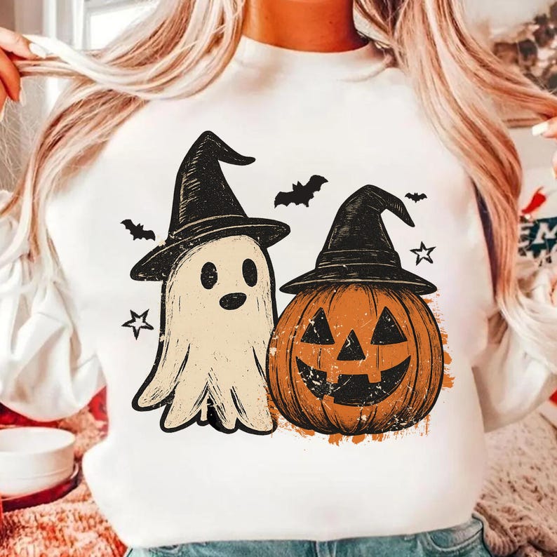 Op de afbeelding: Wit sweatshirt met een Halloween-ontwerp. Het ontwerp bevat een spook met een heksenhoed, een pompoen met een heksenhoed, vleermuizen en sterren. Het spook is gebroken wit en de pompoen is oranje.
