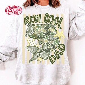 May include: A light gray sweatshirt with a graphic of a fish in camouflage pattern, the words "REEL COOL DAD" in green, and yellow star accents. The sweatshirt has a crew neck.