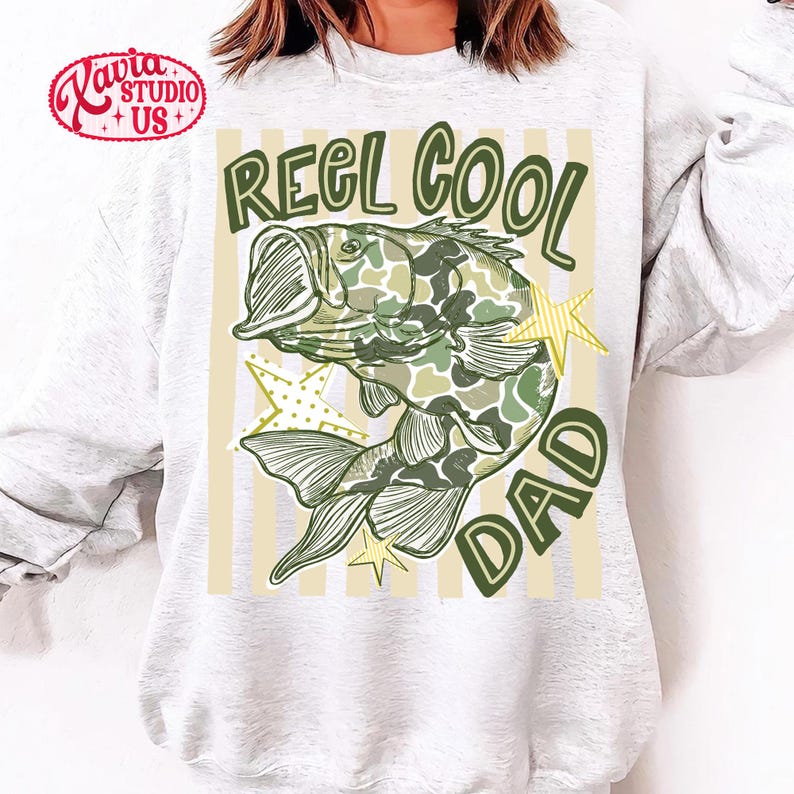 May include: A light gray sweatshirt with a graphic of a fish in camouflage pattern, the words "REEL COOL DAD" in green, and yellow star accents. The sweatshirt has a crew neck.