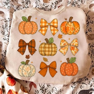 Puede incluir: Sudadera beige con un diseño de calabazas y lazos en tonos naranja, marrón y crema. Las calabazas tienen varios diseños, incluyendo lunares y cuadros. Los lazos son de varios patrones y colores, con pequeñas hojas y estrellas dispersas.