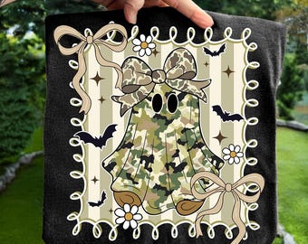 May include: A black square blanket featuring a cartoon ghost in camouflage print, adorned with a large bow. The design includes bats, daisies, and ribbons, set against a striped background. The blanket is held up by a hand.