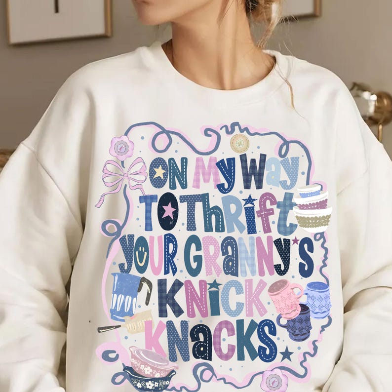 Peut inclure: Sweat-shirt cr&egrave;me avec un motif color&eacute; indiquant &laquo; On My Way To Thrift Your Granny's Knick Knacks &raquo;. Le motif comprend des illustrations de tasses, de bols et d'&eacute;toiles, avec des n&oelig;uds et des motifs floraux.
