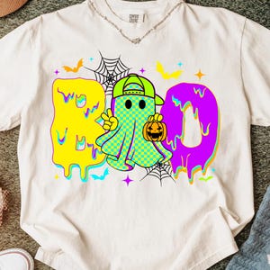 Puede incluir: Camiseta blanca con un colorido gr&aacute;fico de Halloween. El dise&ntilde;o presenta la palabra "BOO" en letras goteantes amarillas y moradas, un personaje fantasma con un signo de la paz, una calabaza y murci&eacute;lagos. La camiseta tiene cuello redondo.