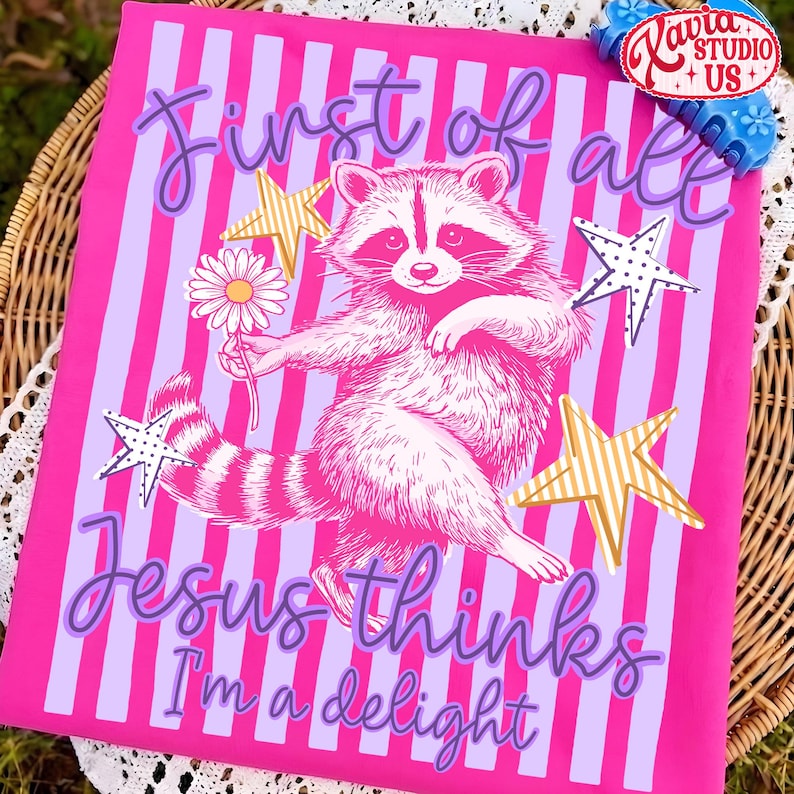 May include: A pink graphic tee with a raccoon illustration holding a daisy, surrounded by stars. The text reads "First of all Jesus thinks I'm a delight" in purple script. The background has vertical stripes.