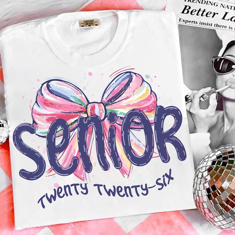May include: White t-shirt featuring a colorful bow design with pink, blue, and yellow hues. The word "Senior" is written in a dark blue font, with "Twenty Twenty-Six" below. A fun and celebratory design.