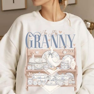 May include: Cream-colored sweatshirt featuring a graphic of a wooden shelf displaying vintage-style dishes, a goose, and a chicken. The text "In My Granny Era" is printed in blue script above the shelf design. The design includes hearts.