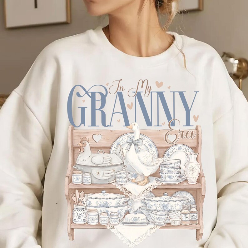 May include: Cream-colored sweatshirt featuring a graphic of a wooden shelf displaying vintage-style dishes, a goose, and a chicken. The text "In My Granny Era" is printed in blue script above the shelf design. The design includes hearts.