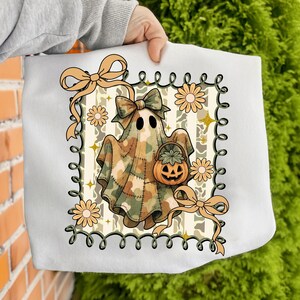May include: White tote bag featuring a Halloween-themed design. The design includes a ghost wearing a camouflage pattern sheet, holding a pumpkin basket. The ghost is surrounded by flowers, bows, and a decorative border. The overall color scheme is beige, brown, and orange.