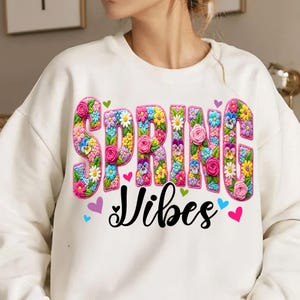 Puede incluir: Sudadera blanca con la frase "SPRING Vibes". La palabra "SPRING" est&aacute; en un dise&ntilde;o floral colorido en 3D. El dise&ntilde;o incluye rosas, margaritas y pensamientos. Peque&ntilde;os corazones multicolores.